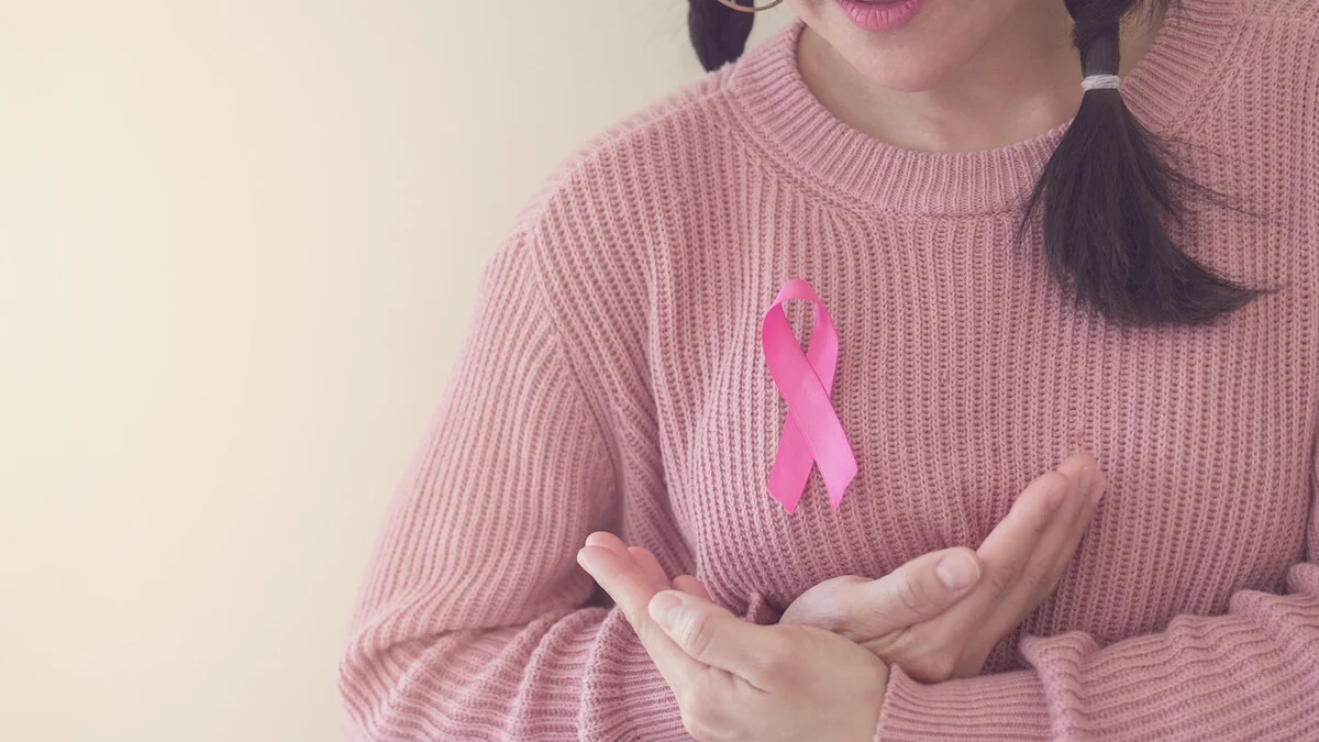Mulher com suéter rosa segurando laço rosa da campanha Outubro Rosa junto ao peito em gesto de autocuidado